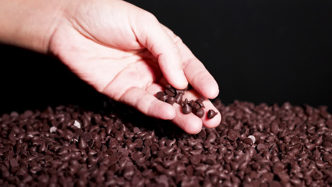 A hand sorts chocolate drops against a dark backdrop, highlighting texture and contrast in a close-up, well-lit setting