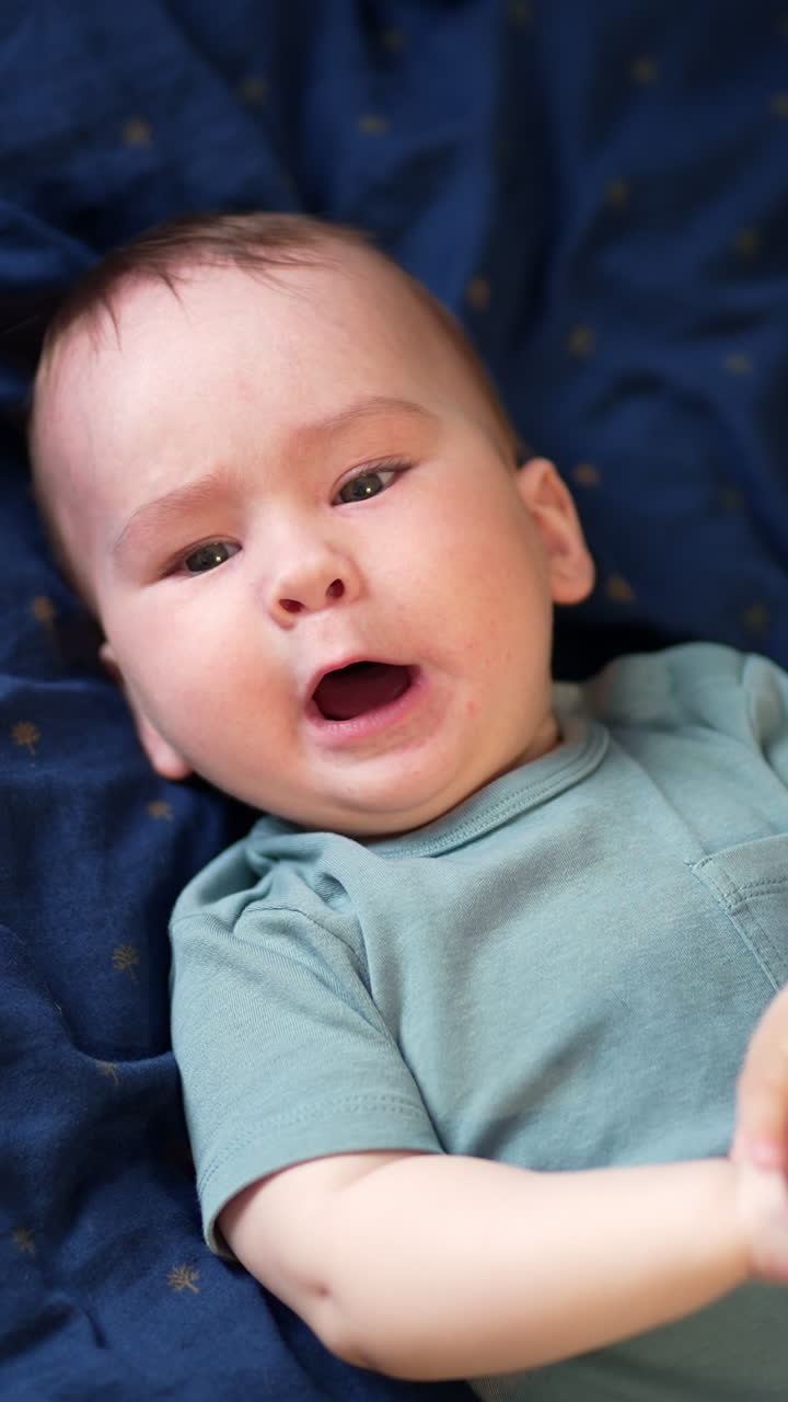 Active Caucasian baby boy lies on his back opening mouth to scream. Cute child waving hands and legs with anxiety. Top view. Vertical video