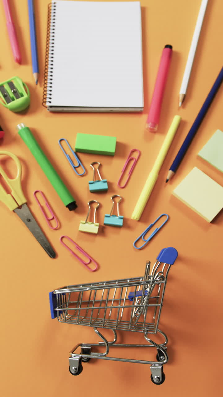 Vertical video of overhead view of shopping trolley and school items on orange background