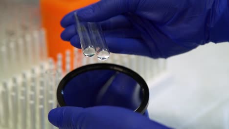 blue latex gloved hands pick up and inspect small test tube vials while holding over a mirror