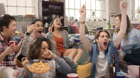 diverse group of student friends arms raised celebrating goal watching sports event on tv together eating snacks  drinking beer