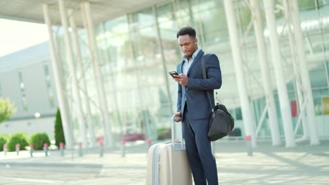 african american business man standing on the background a modern train station airport in formal suit with a suitcase using app mobile phone.