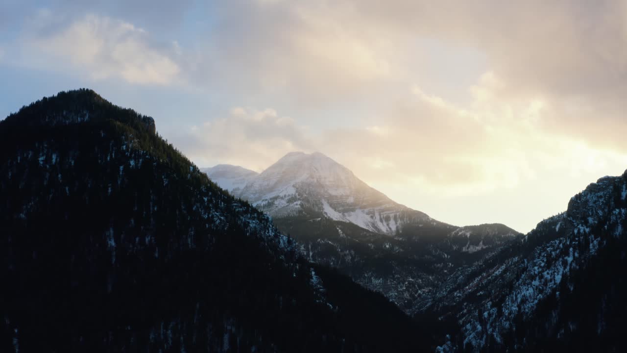 Descending aerial drone shot of a winter landscape of Mount Timpanogos in the background surrounded by a pine tree forest during sunset from Tibble Fork Reservoir in American Fork Canyon, Utah
