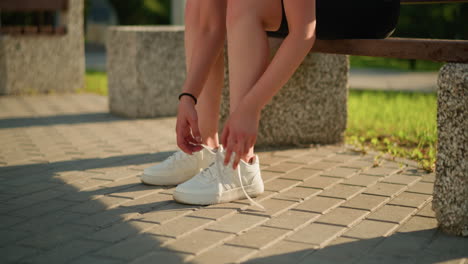 partial view of lady seated on bench outdoors removing left sneaker with left hands,while her right hand is on the leg sunlight casting shadows on paved ground, greenery in background