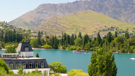 Handheld wide shot over Lake Wakatipu on New Zealand’s South Island showing a ship crossing calm waters with a distant mountain backdrop. Parts of Queenstown can be seen to the left