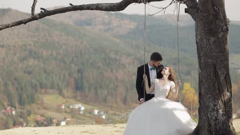 newlyweds. caucasian groom with bride ride a rope swing on a mountain slope