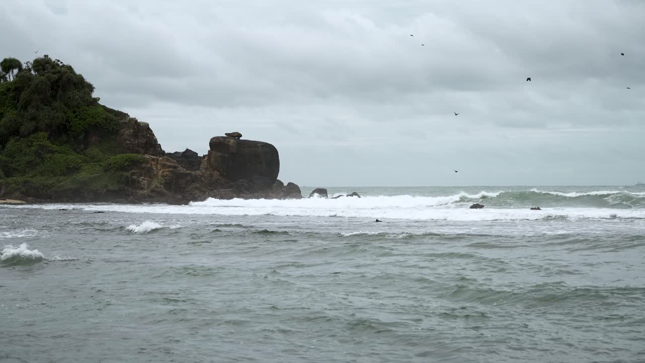 스리랑카의 바위 위에 부히는 바다 파도, 위에서 날아다니는 새, 배경에는 구름이 가득한 하늘