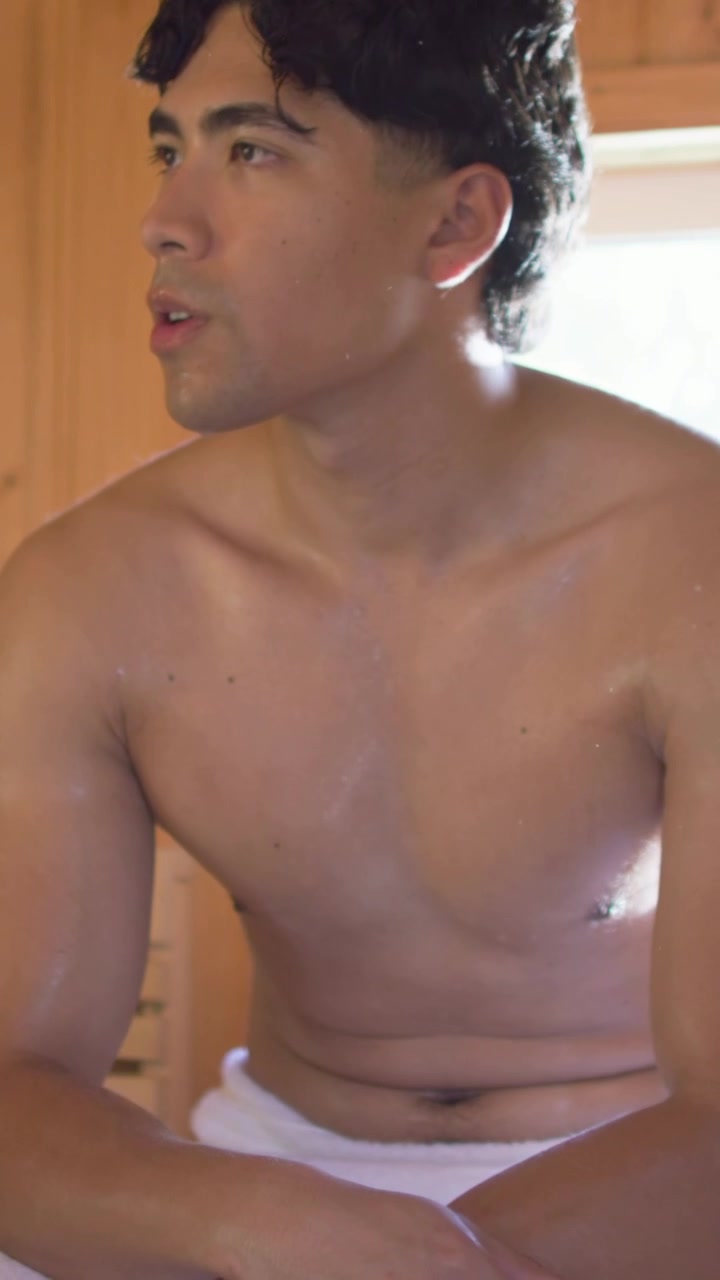 Video verticale: Luce solare che riscalda un uomo che sposta lo sguardo e appoggia le mani su un asciugamano in una sauna vicino al vetro.