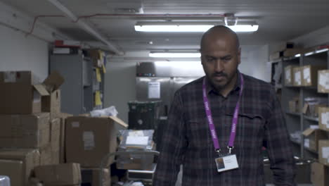 bald indian male walking towards camera in office basement and stopping to scan shelves for item