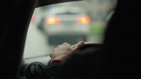 back view of gentleman driving with hands on steering wheel, blurry parked car visible ahead through rain speckled windshield, focus on hand grip and road ambiance in urban setting during commute