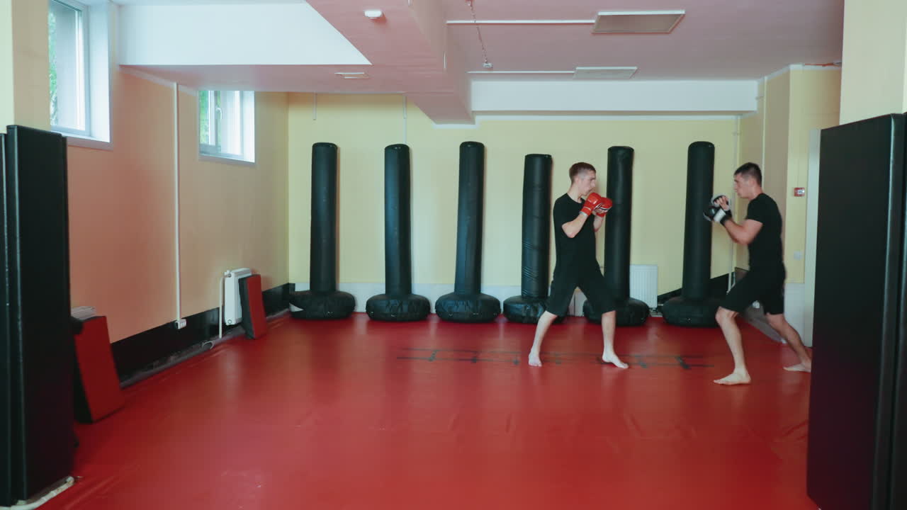 Atletas practicando sparring en un gimnasio de artes marciales; uno de ellos da un puñetazo mientras el otro bloquea en el suelo de la colchoneta roja. Sesión de entrenamiento rodeada de sacos de boxeo verticales en un ambiente de lucha competitiva.