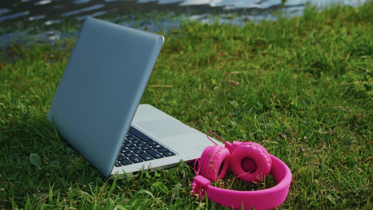 Laptop e cuffie rosa poggiano sull'erba vicino all'acqua. Atmosfera di spazio di lavoro all'aperto pacifico. Ideale per lavoro da remoto, studio o progetti creativi. La luce naturale migliora la calma e la concentrazione.
