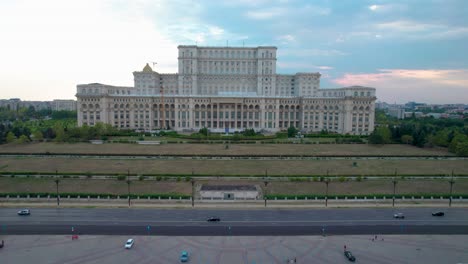 Golden-Hour-Aerial-View-of-the-Palace-of-Parliament-in-Bucharest,-Romania,-with-Lush-Vegetation-and-Glistening-Water-Fountains,-Backward-Movement