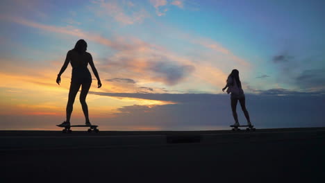 Amid-the-enchanting-hues-of-a-sunset,-two-friends-donning-shorts-engage-in-slow-motion-skateboarding-on-a-road.-Mountains-and-a-scenic-sky-provide-the-setting