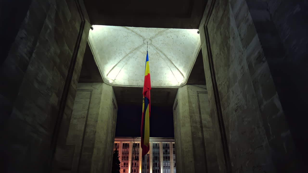 A large Venezuelan flag waves in the night sky, illuminated by the lights of nearby buildings. The flag stands out against the concrete structure, showcasing national pride