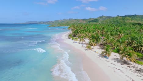 Cinematic-drone-flight-over-sandy-beach-with-palm-trees-and-turquoise-Caribbean-sea-water---Mountain-landscape-in-background---Playa-Rincon,-Samana