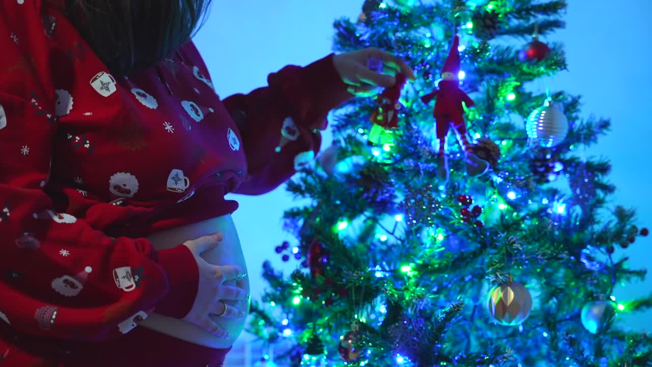 Medium close up shot of a pregnant woman in red pajamas uncovering her belly to touch it in front of the lights of the Christmas tree