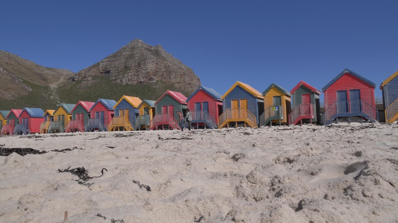 Dos mujeres caminan por la playa pasando las icónicas, vibrantes y multicolores casetas de playa en la playa de Muizenberg, Ciudad del Cabo, con una escarpada cordillera visible a lo lejos bajo un cielo azul claro