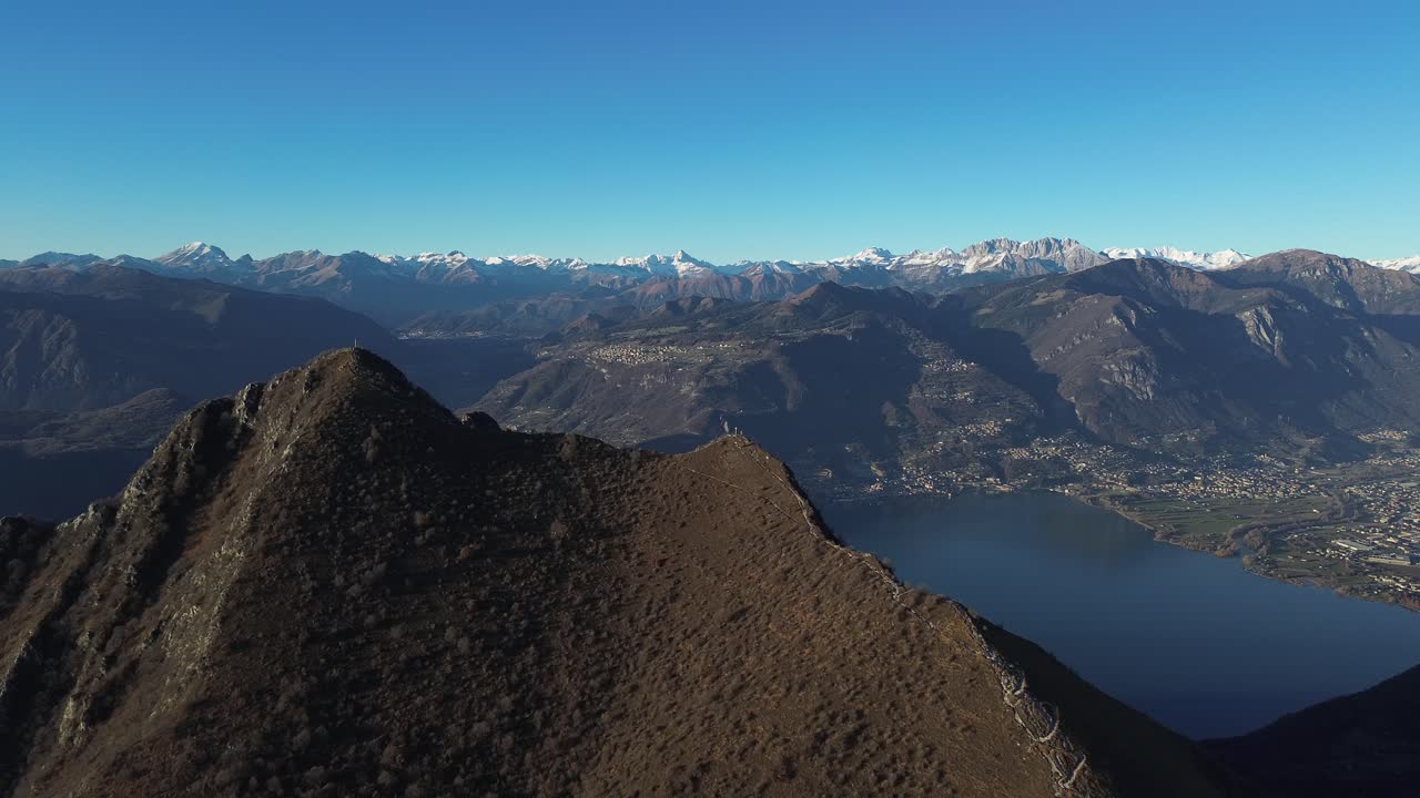 Drone shot over Lake Iseo with surrounding mountains in Italy and snow-covered peaks visible far away.