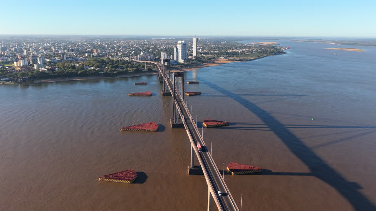 Rastreamento de drone em alta altitude à esquerda, angulado levemente para baixo para mostrar caminhões se movendo sobre a ponte e a água marrom turva do rio abaixo