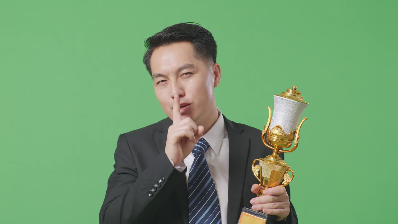 슈트와 타이를 입은 아시아 사업가, 금메달과 트로피를 들고, 조용한 제스처를 하고, 그라운드에서 초록색 스크린에서 첫 번째 우승자로서 승리하는 것이 비밀이라고 말합니다.