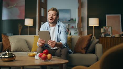 Hombre-Amigable-Agitando-La-Mano-Hablando-Con-Una-Tableta-En-Casa.-Chico-Relajado-Sentado