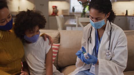 Mixed-race-female-doctor-wearing-face-mask-filling-syringe-at-home