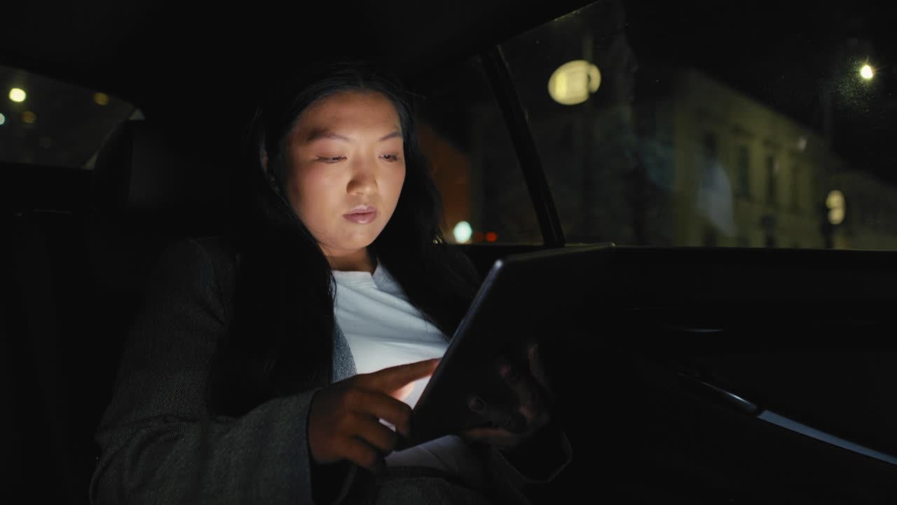 비즈니스 중국 여성이 택시를 운전하고 밤에 디지털 태블릿을 탐색하고 있습니다.
