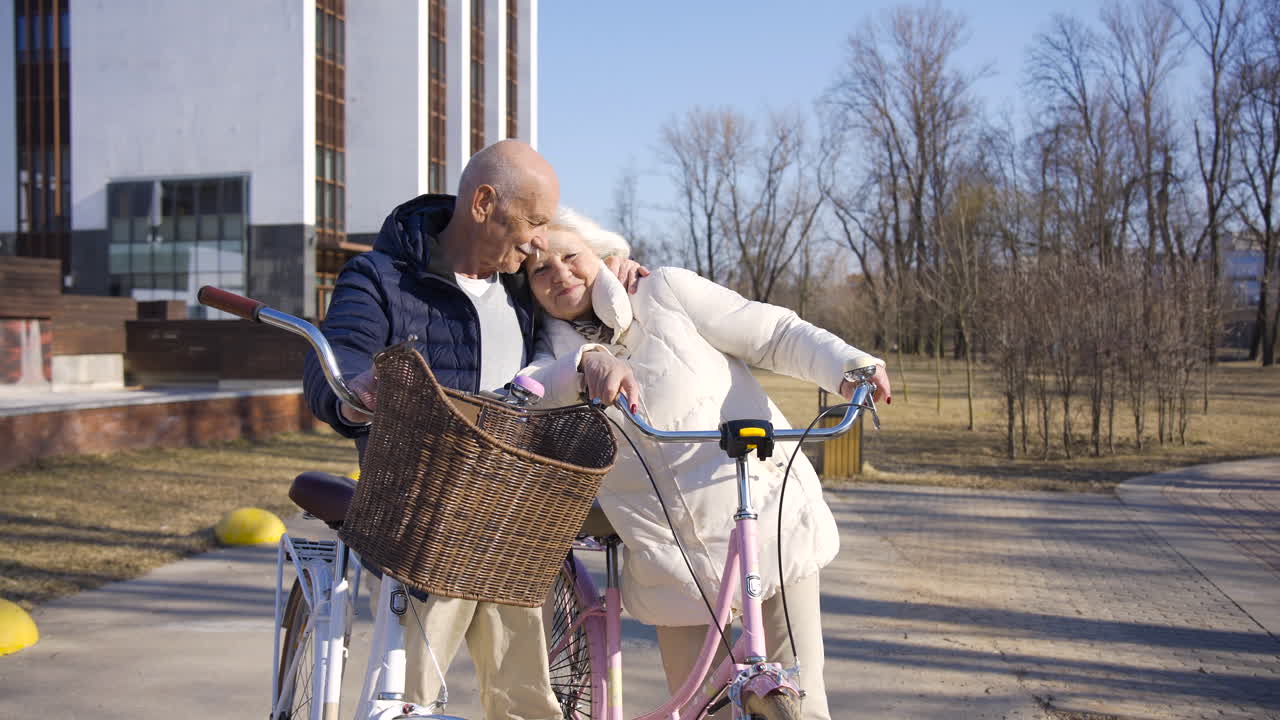 pareja mayor sosteniendo bicicletas mientras se miran, abrazándose y sonriendo en el parque en un día de invierno