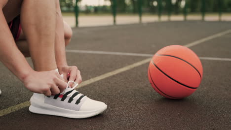 Primer-Plano-De-Un-Jugador-De-Baloncesto-Masculino-Poniéndose-El-Zapato,-Atando-Los-Cordones-De-Los-Zapatos-Y-Luego-Recogiendo-La-Pelota-Del-Suelo-En-Una-Cancha-De-Baloncesto-Al-Aire-Libre