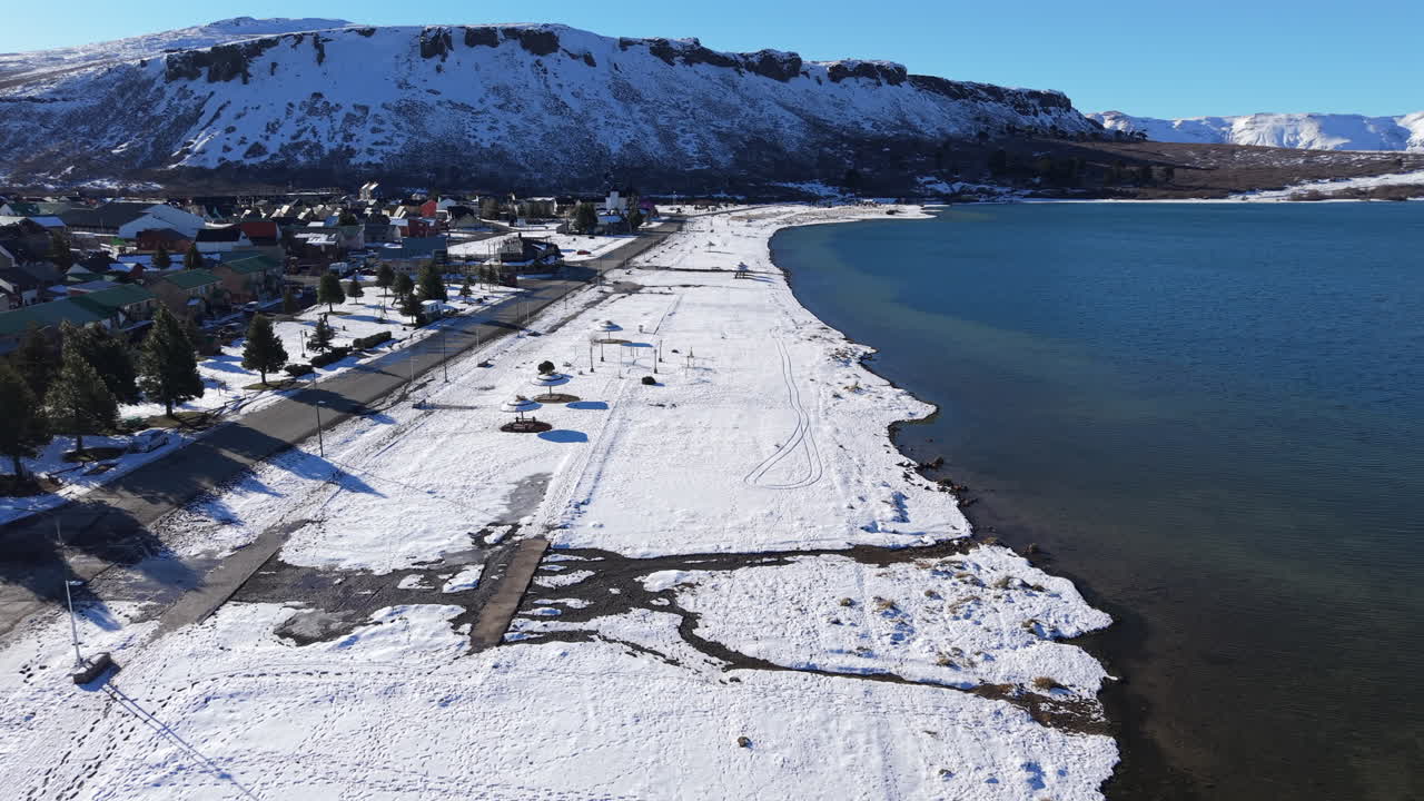Drone glides down toward the icy shores of Lake Caviahue, revealing the volcanic slopes of Volcán Copahue and the surrounding Patagonian Andes under a pristine winter blanket