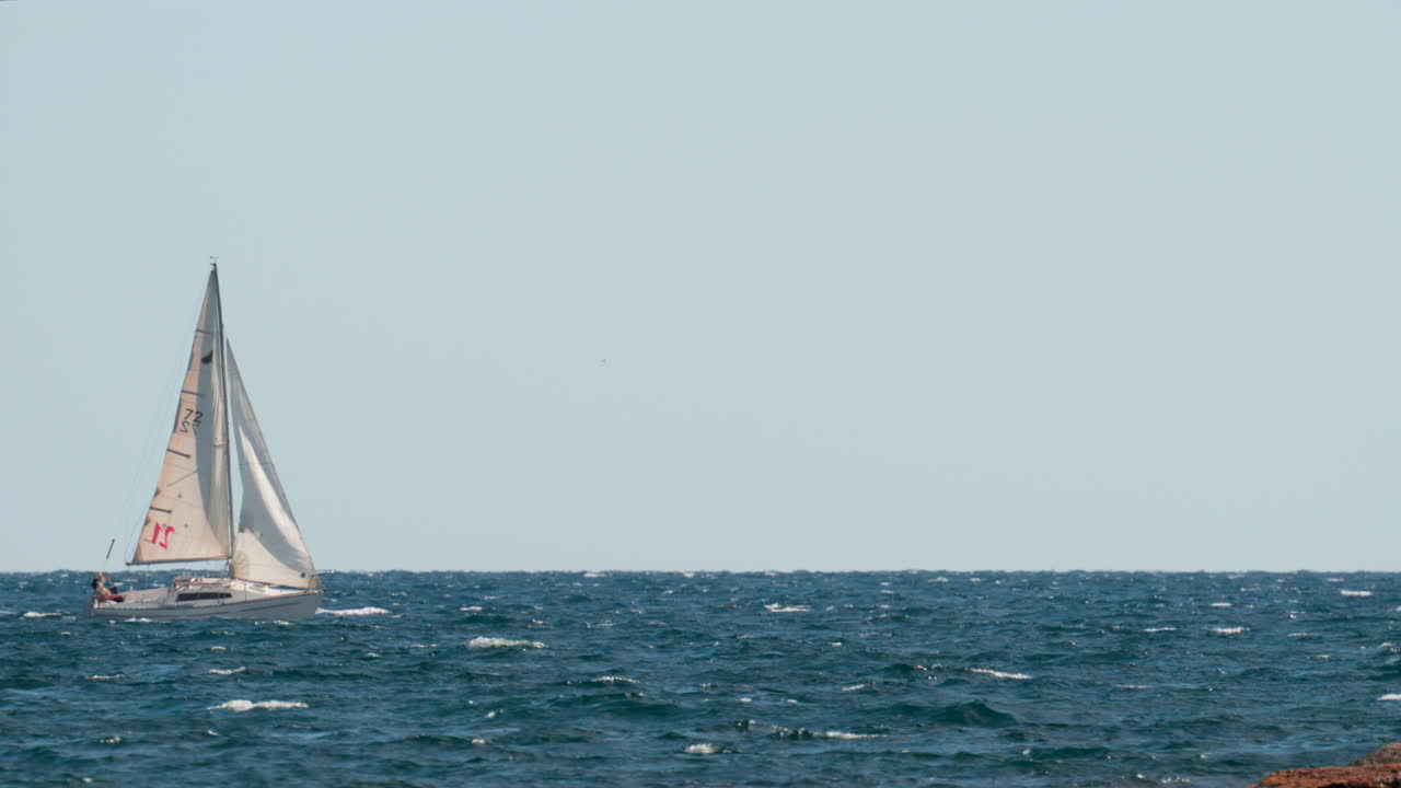 Cannes, France - October 7, 2025: A white sailboat navigates through blue waves on a clear sunny day, with the horizon stretching endlessly behind it