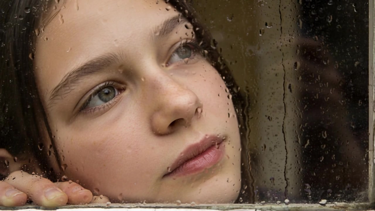Following raindrops, teen tilting and tracking them at home window, thinking, hand resting on sill