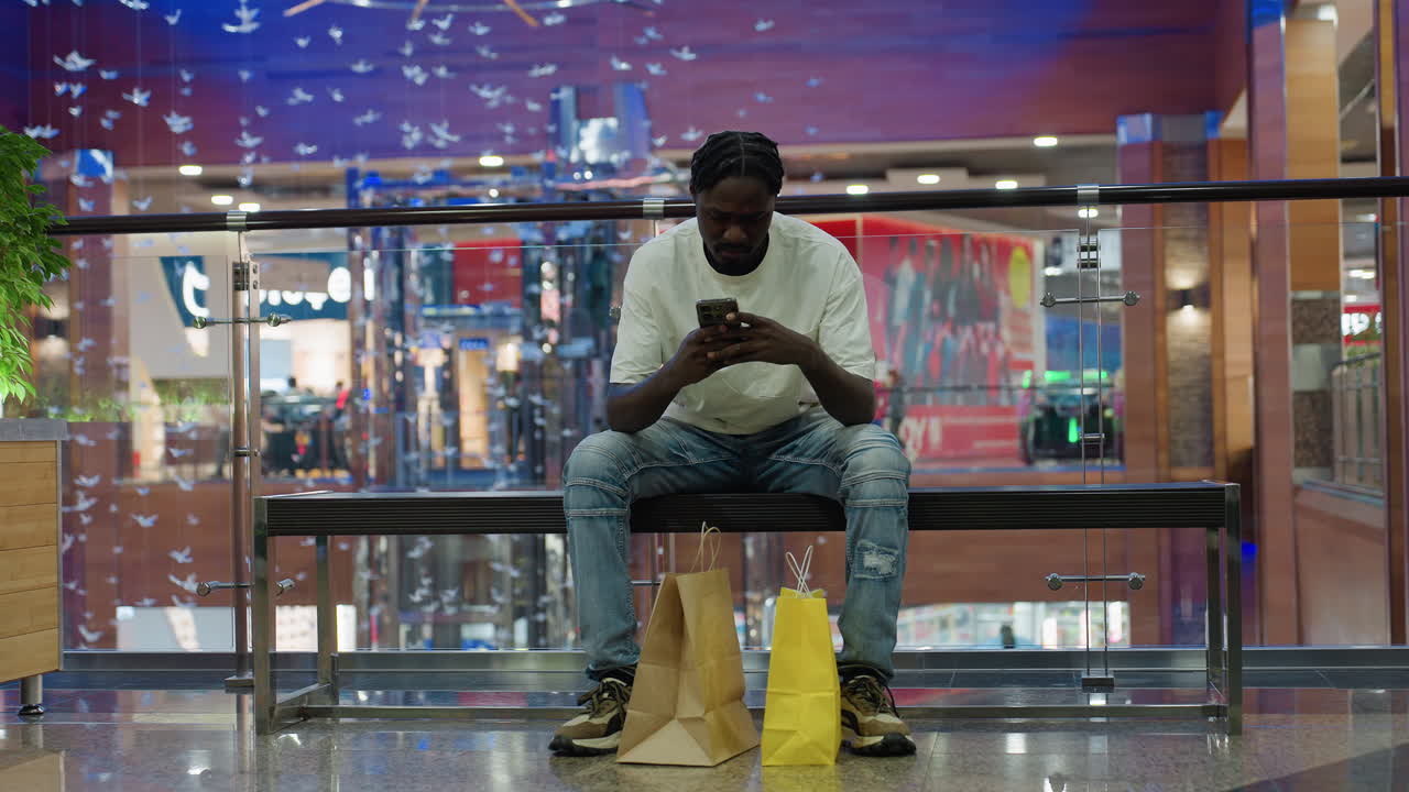 Young black man in white shirt and jeans sits on bench inside bright shopping mall focused on smartphone, surrounded by shopping bags, decorative plants, hanging glass birds, and blurred shoppers
