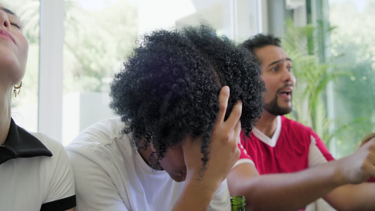 Diverse friends watching game leaning forward grimacing then calming red jersey with beer on couch