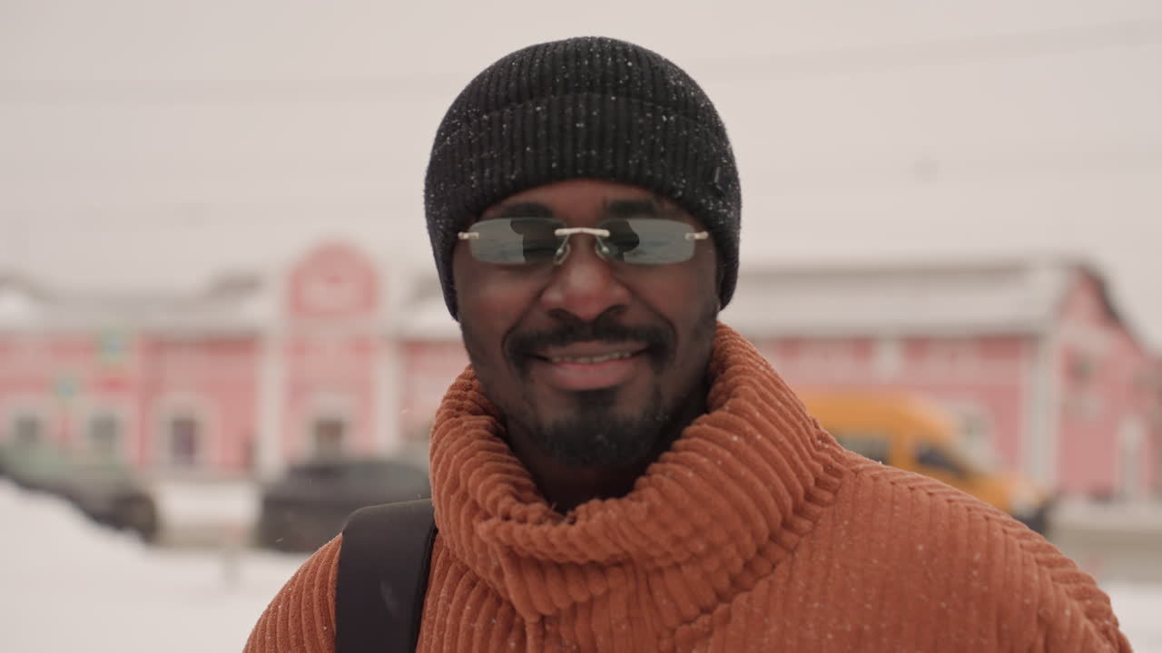 Uomo di colore in una strada innevata, che indossa un maglione di lana arancione e un berretto nero, parla al telefono sorridendo, con una vetrina rosa pastello e lampioni vintage sullo sfondo, leggera nevicata, pendolare.