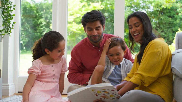 Smiling Family At Home Sitting On Floor In Lounge Reading Book Together