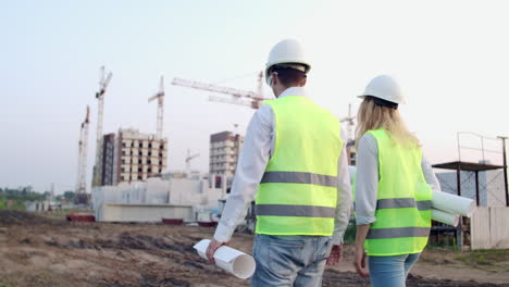 Back-turned-engineer-and-controller-at-construction-site-reviewing-office-blueprints-and-technical-drawings-walk-and-discussing.