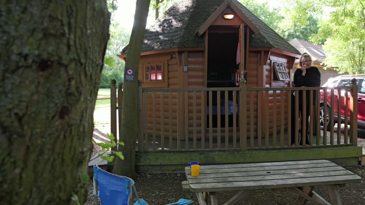 Reveal shot of woman talking on phone while standing on porch of cozy wooden cabin in forest setting.