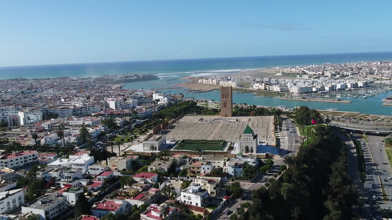 Toma de dron ultra amplia que captura la extensión completa de Rabat. Presenta el histórico complejo de la Torre Hassan, el moderno Puente VI que cruza el Bou Regreg y las ciudades que se encuentran con el Océano Atlántico.