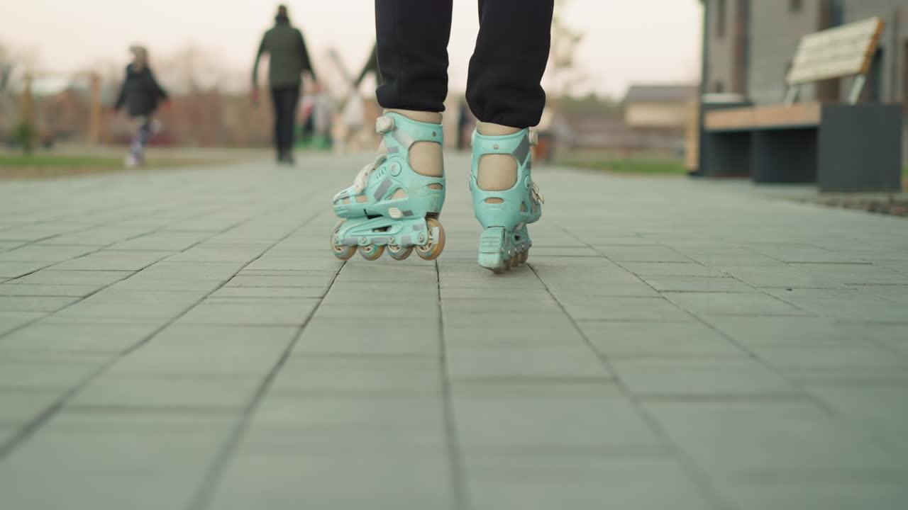 A close-up shot of rollerblades in motion, gliding through a paved park path. The background is blurred, featuring people and benches, capturing the dynamic movement and urban park environment