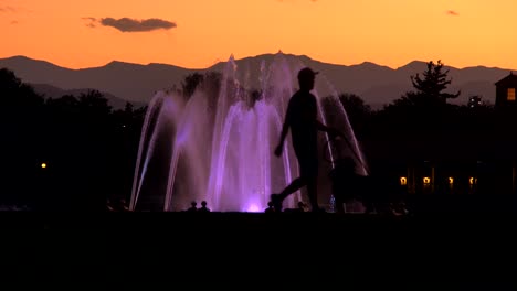 Hombre-Paseando-A-Un-Perro-Sobre-Un-Fondo-De-Fuente-De-Agua-Iluminada-Y-El-Horizonte-De-Denver-Al-Atardecer