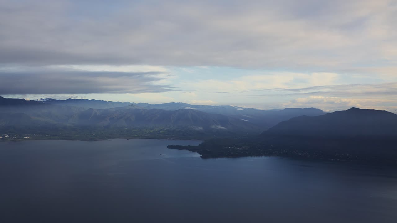 Et luftfoto fra et flyvindue af en massiv tropisk ø, omgivet af et tågeslør og spredte skyer i de tidlige morgentimer.