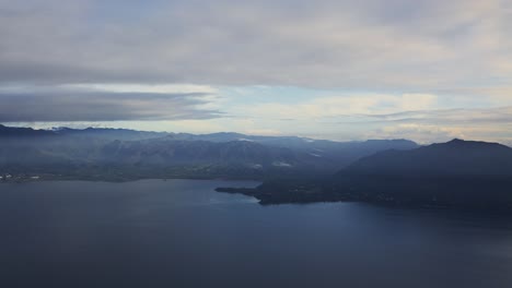 Eine-Luftaufnahme-Aus-Dem-Fenster-Eines-Flugzeugs-Einer-Riesigen-Tropischen-Insel,-Die-In-Den-Frühen-Morgenstunden-Von-Einem-Nebelschleier-Und-Vereinzelten-Wolken-Umgeben-Ist