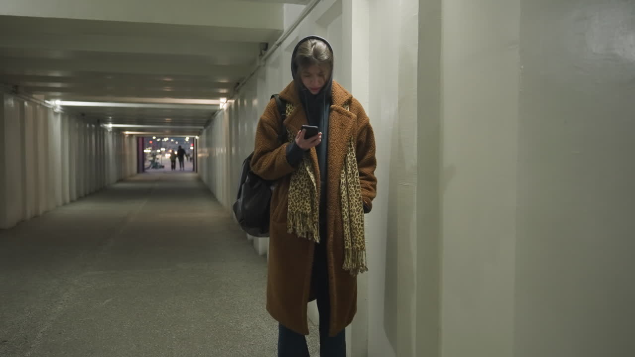 A girl in a brown coat and jeans walks through an underpass tunnel, leaning against the wall while looking at her phone with a worried and sad expression. a moment of anxiety and loneliness