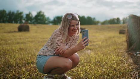 woman squatting in golden field, smiling warmly as she takes selfie with smartphone, sunlight casts a soft glow on her face, capturing joyful moment outdoors in peaceful countryside setting