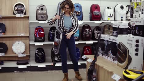 cheerful curly haired woman standing near new vacuum cleaners in home appliance store, choosing the right one. various vacuum