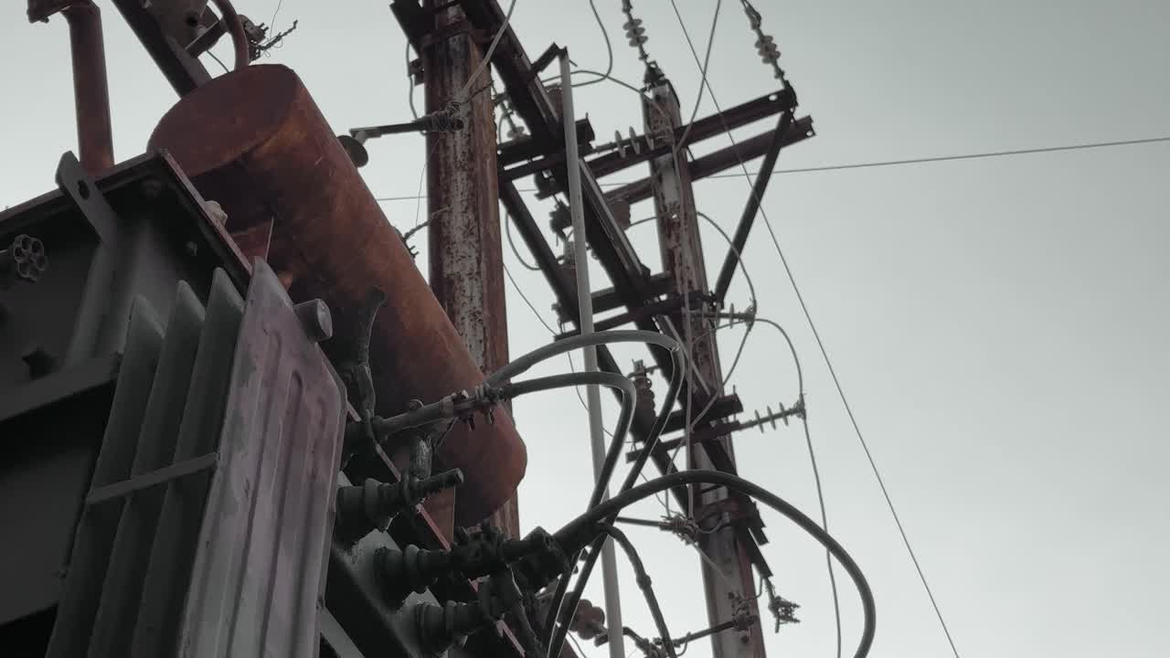 A tilt-down shot of an electrical transformer connected to overhead power lines on a rusted pole. The camera pans downward, revealing the complex network of wires and insulators