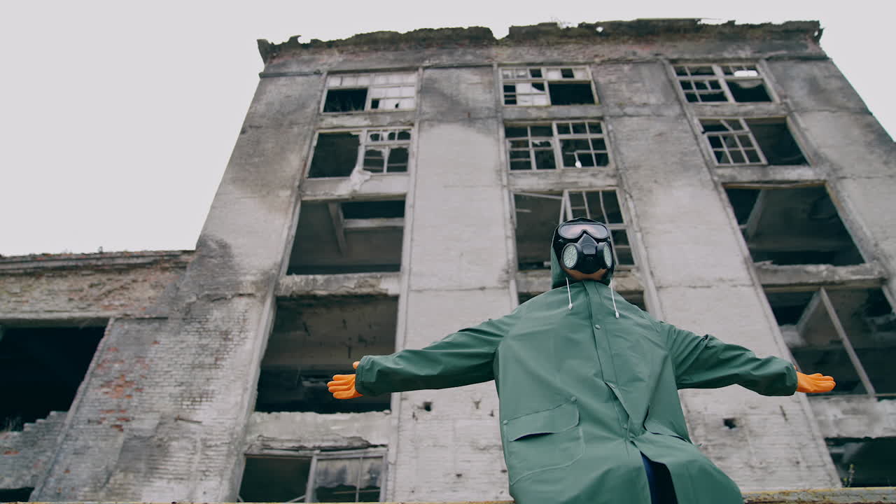 Man with gas mask in ruined place. Human in protective suit and respirator sitting outdoors with outstretched arms on the background of destroyed building. Radioactive zone.