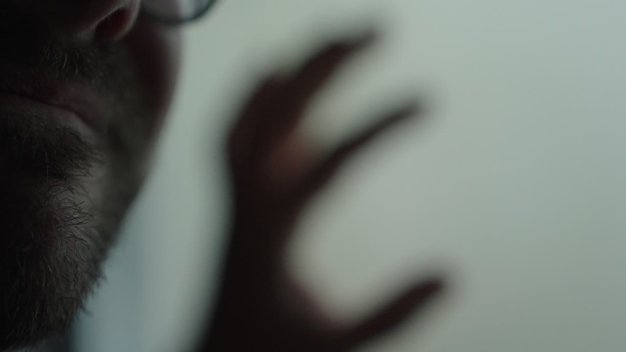 Primo piano introspettivo di un uomo barbuto in condizioni di scarsa illuminazione con un gesto della mano sfocato, che esprime disperazione e lotta emotiva. Ideale per temi come depressione, ansia, crisi, dolore e salute mentale drammatica.